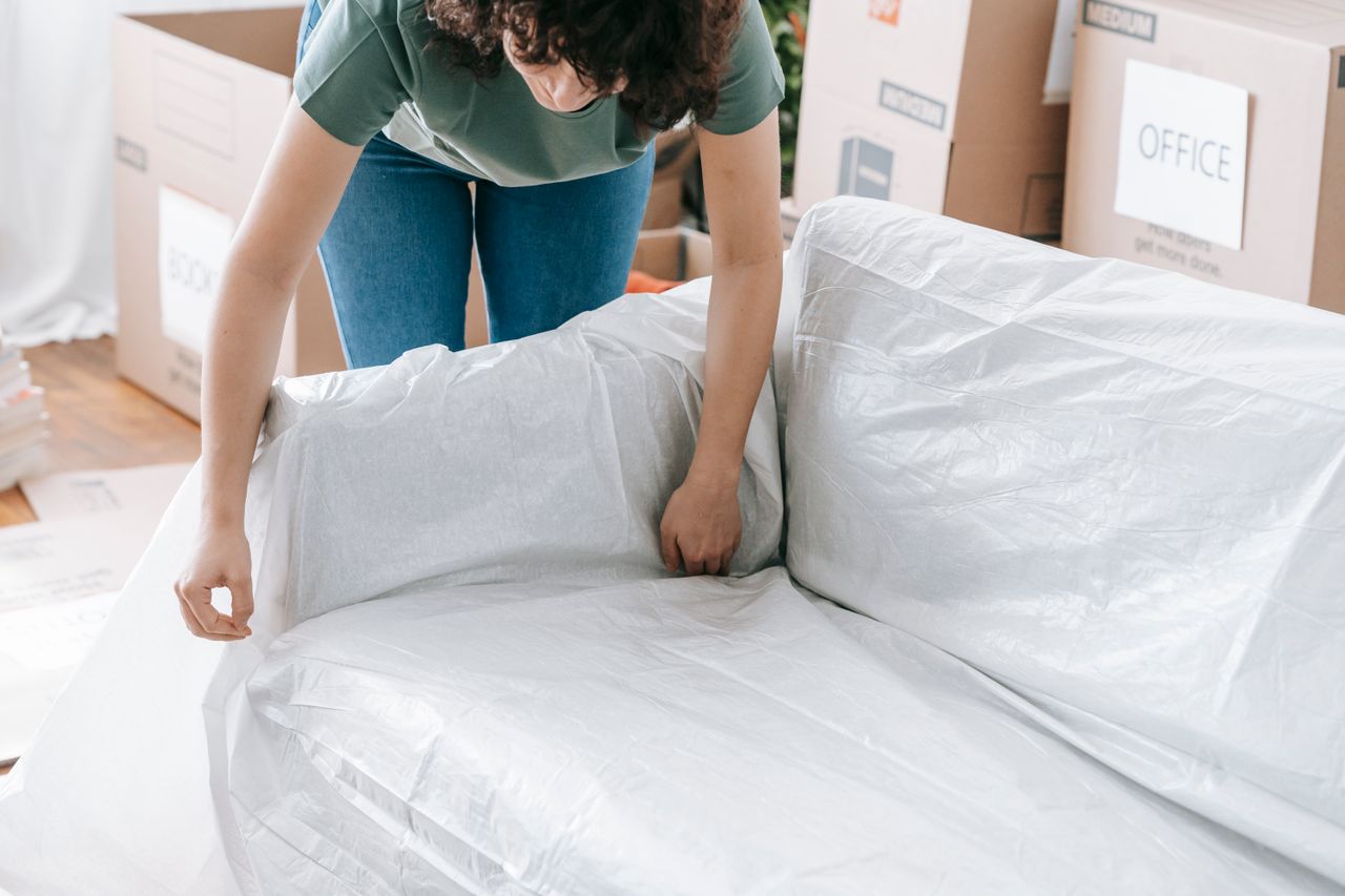 A woman gently covers a sofa with protective wrapping while packing boxes in a bright room.